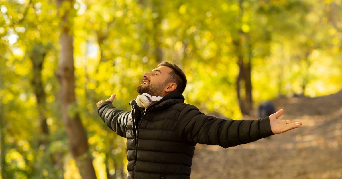 A person in a black jacket stands in a Utah forest, arms outstretched and wearing headphones around their neck, enjoying the sunlight filtering through the trees—perhaps finding solace in nature after seeking treatment services for alcohol & drug abuse.