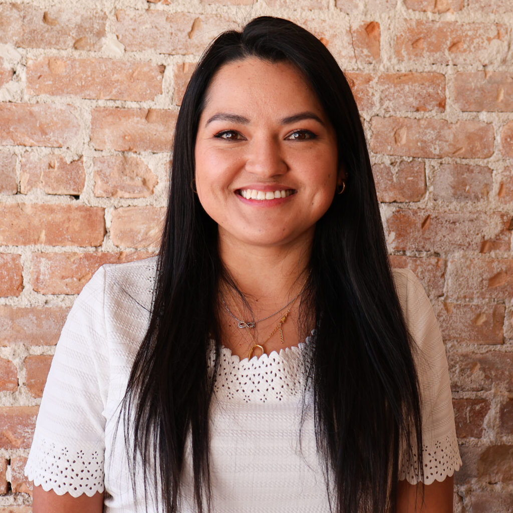 Woman with long black hair and a white top smiles in front of a brick wall.
