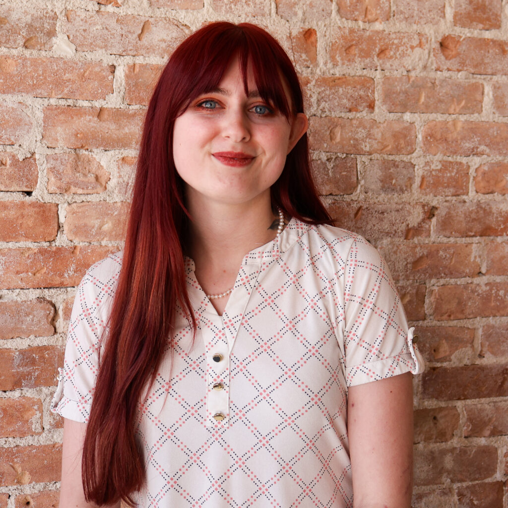 Woman with long red hair wearing a patterned white blouse stands in front of a brick wall, smiling.