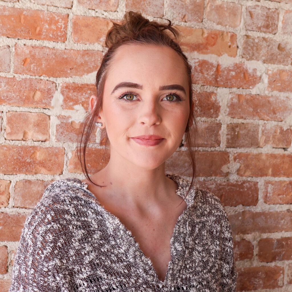 A person with brown hair in a bun, wearing a gray knitted sweater, stands in front of a brick wall.