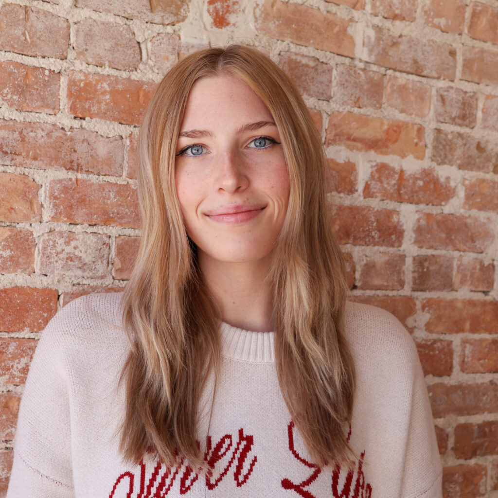 Client Outcomes A person with long blonde hair and a neutral expression stands against a brick wall, wearing a sweater.