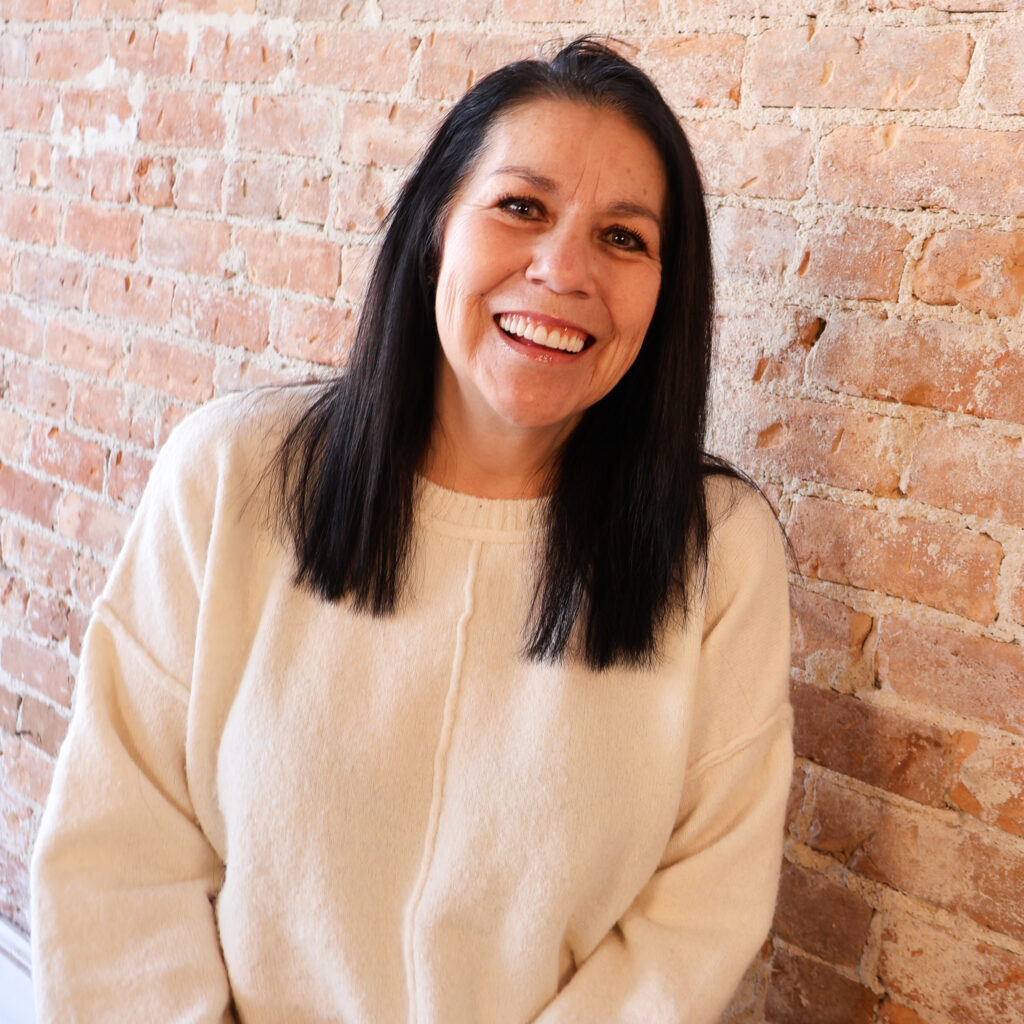 Person with long dark hair smiling, wearing a cream sweater, standing against a brick wall.