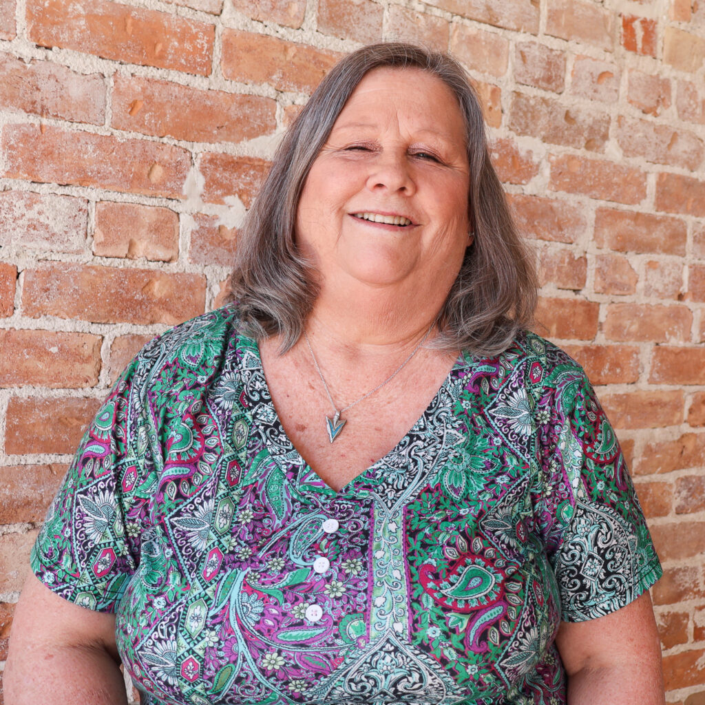 A woman with shoulder-length gray hair smiles while standing against a brick wall. She is wearing a colorful paisley patterned blouse.