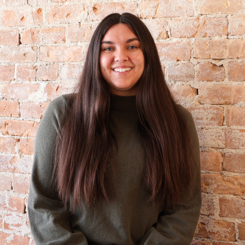 A person with long brown hair and a green sweater is smiling in front of a brick wall.